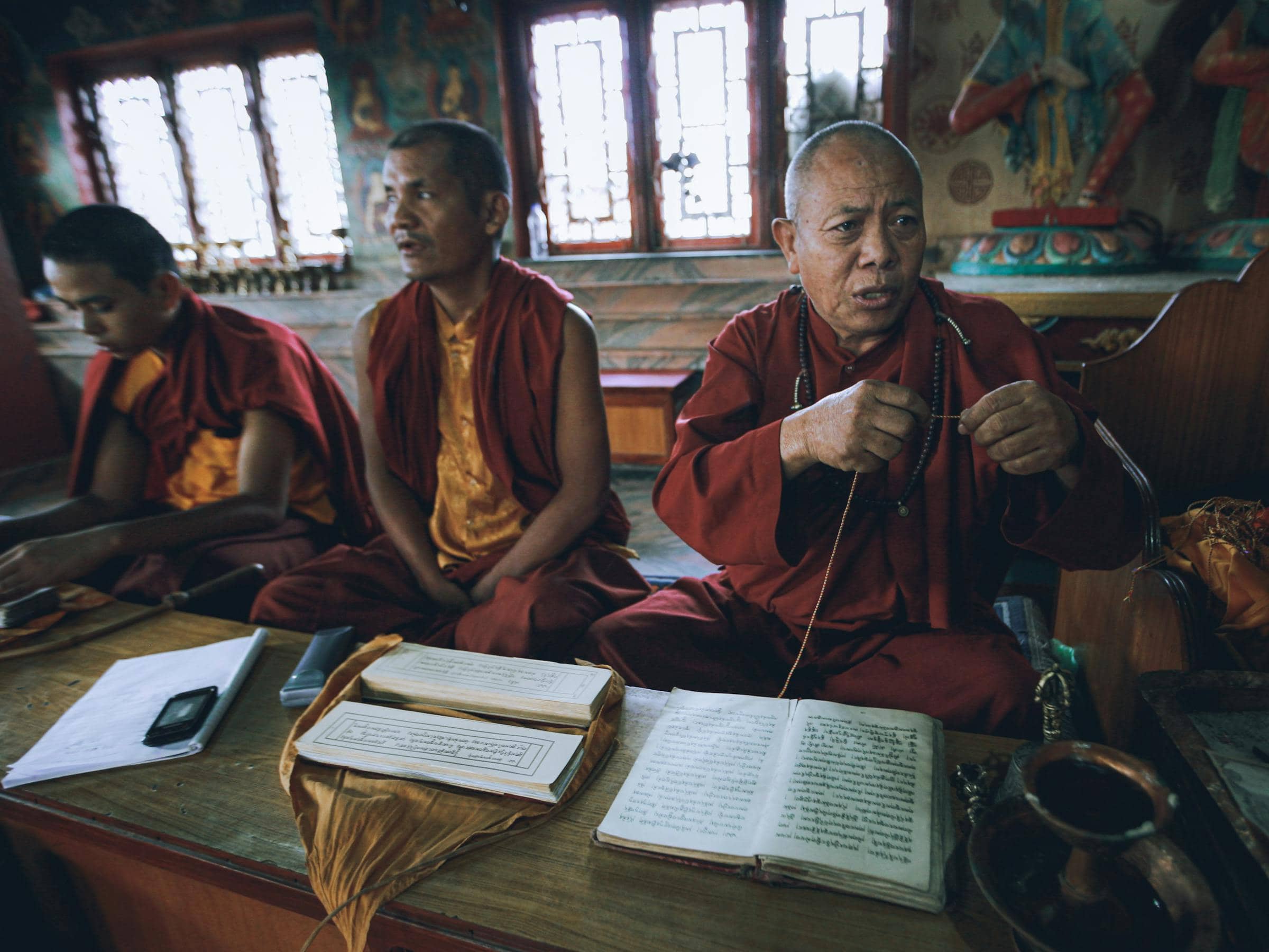 Three Buddhist monks reading scriptures with prayer beads inside a candlelit monastery hall.